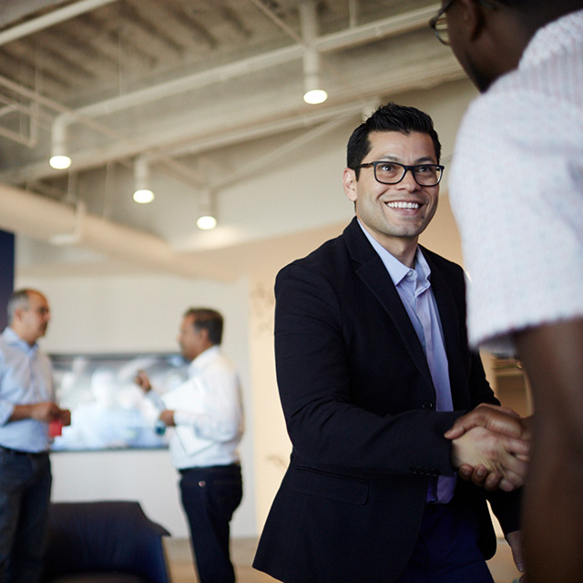Two men shaking hands while two more are talking with each other in the background.